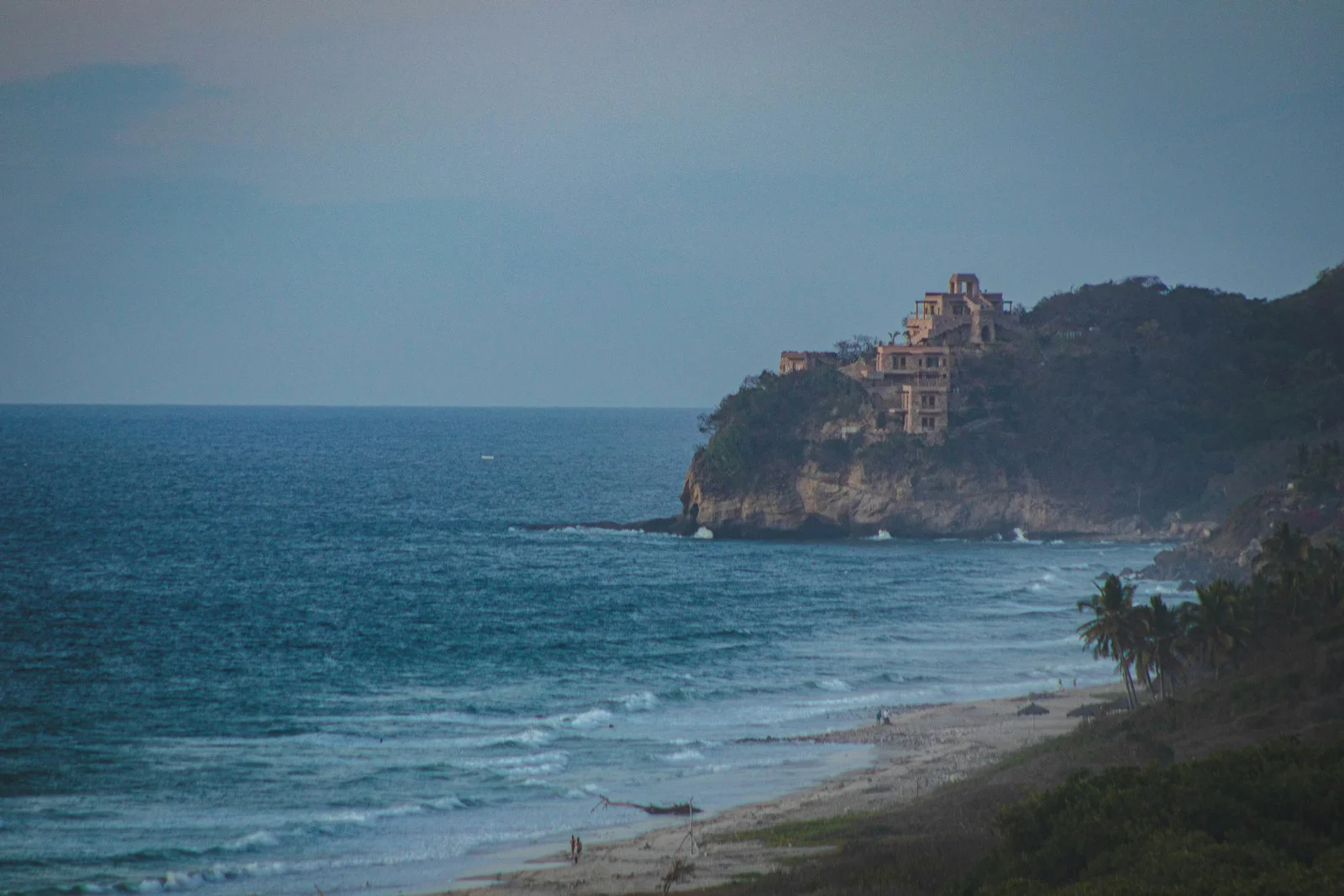 Rocky coastal cliff at Punta de Mita seen from the sea, with a villa on the point and palm trees along the beach